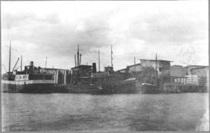 Kangassaaren sahan ranta veden yli joskus 1922-37. Laiturissa höyrylotja (tervahöyry) s/s Puuliike (kotipaikka Viipuri), hinaaja s/s Täljsten ja tunnistamaton apuhinaaja. Sahan rakennuksia, sahatavarataapeleita.