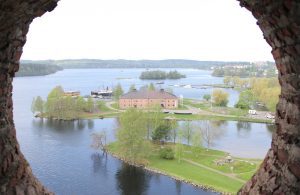 Kuvassa otettu valokuva on otettu Olavinlinnan tornin pyöreän ikkuna-aukon kautta siten, että linnanmuuri kehystää kuvasta aukeavaa maisemaa. Maisemassa näkyy Riihisaaren museo ja sen laiturissa olevat museolaivat sekä taustalla avautuva Pihlajavesi.