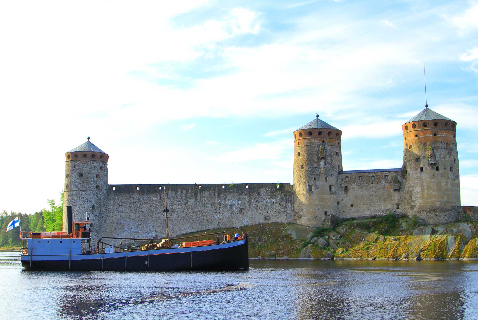 Mikko-laiva ohittamassa Olavinlinnaa. Kuvassa Mikko-laiva purjehtii kesäisessä maisemassa Savonlinnan kaupungin vesillä. Kuvassa taustalla näkyy Olavinlinna.