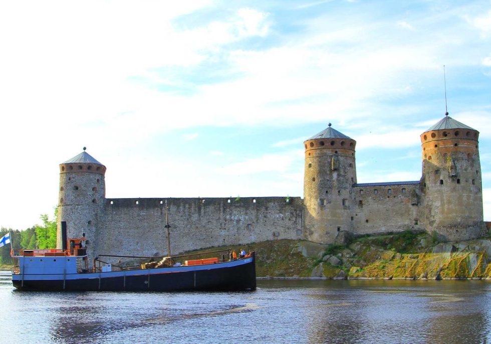 Kuvassa Mikko-laiva purjehtii kesäisessä maisemassa Savonlinnan kaupungin vesillä. Kuvassa taustalla näkyy Olavinlinna.